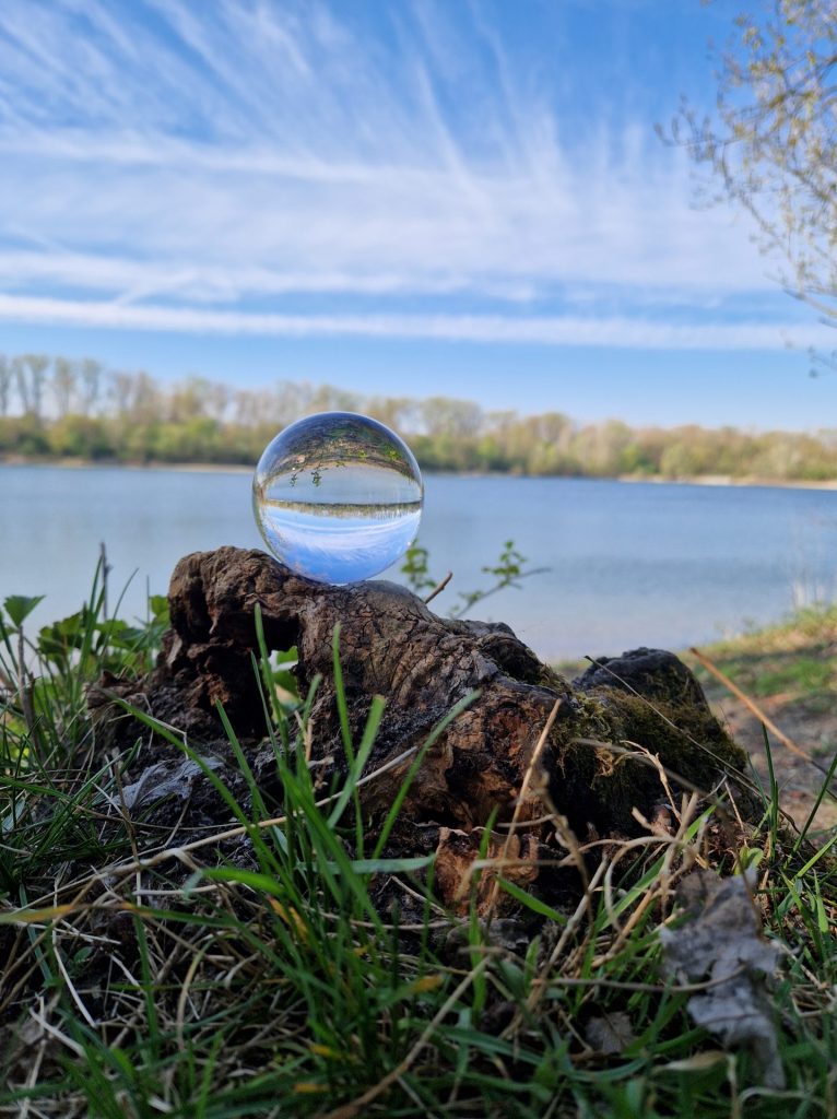 Eine Glaskugel, Perspektivenwechsel, im HIntergrund des sees und die Kugel ist auf einer Wurzel abgelegt 